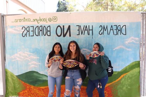 三个学生在“梦想无国界”壁画前用纸蝴蝶庆祝节日